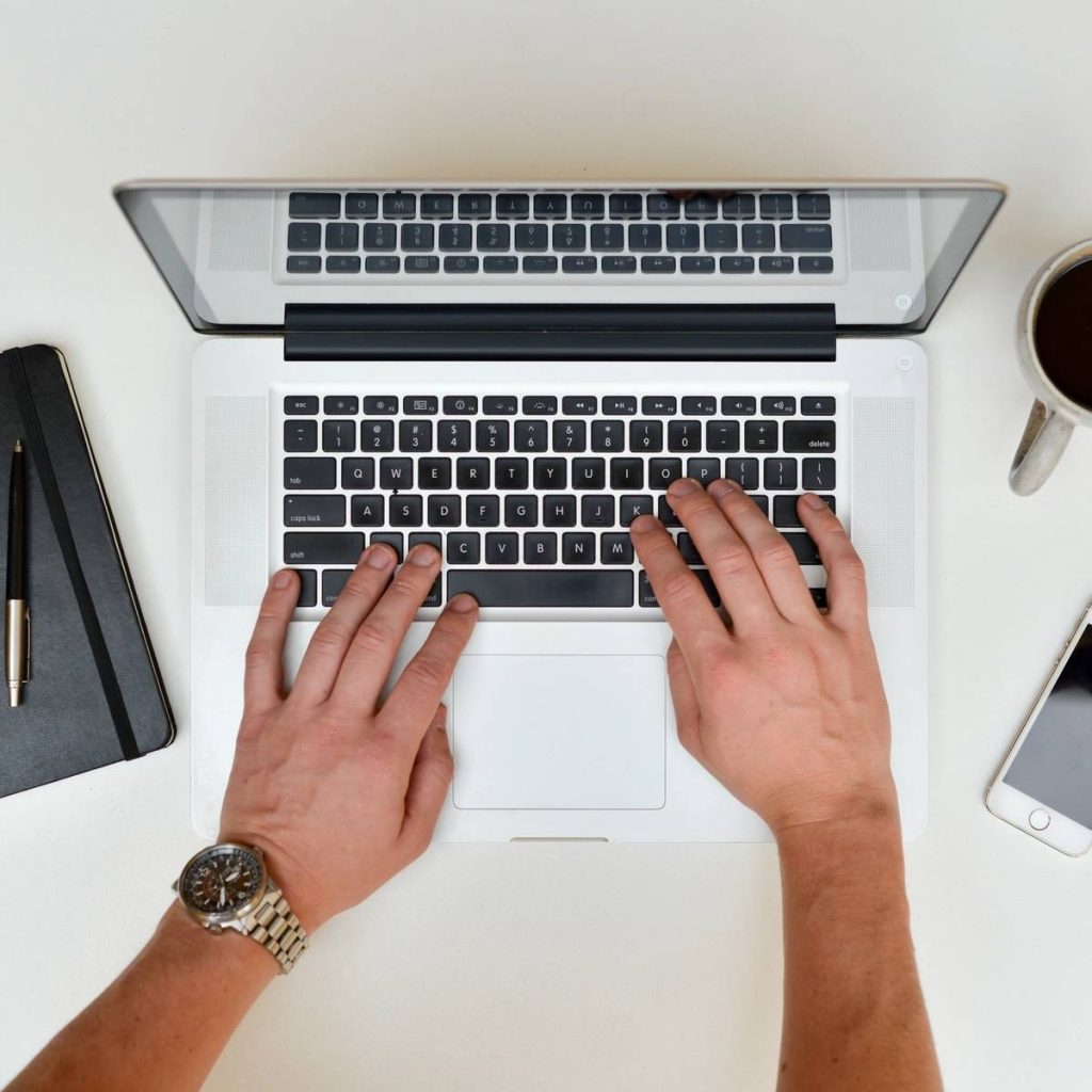 Top view of a person engaged in online learning, typing on a laptop keyboard. A notebook with a pen is on the left, a cup of coffee on the right, and a smartphone next to it. The watch-clad left wrist adds style while delving into digital marketing strategies.