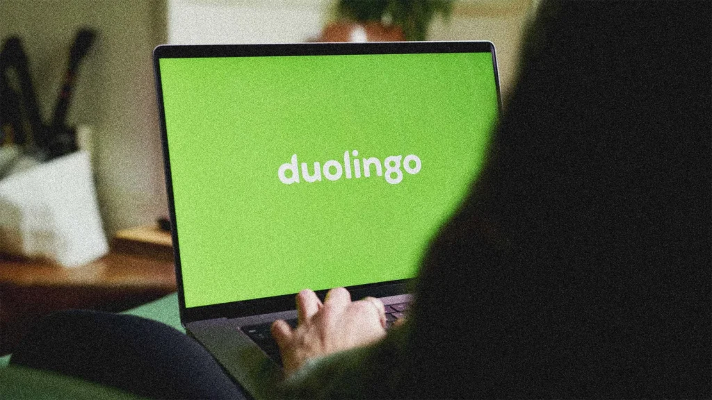 A person uses a laptop displaying the Duolingo logo on a bright green screen. The individual is seated indoors with their hand on the laptop keyboard. Some objects are blurred in the background.