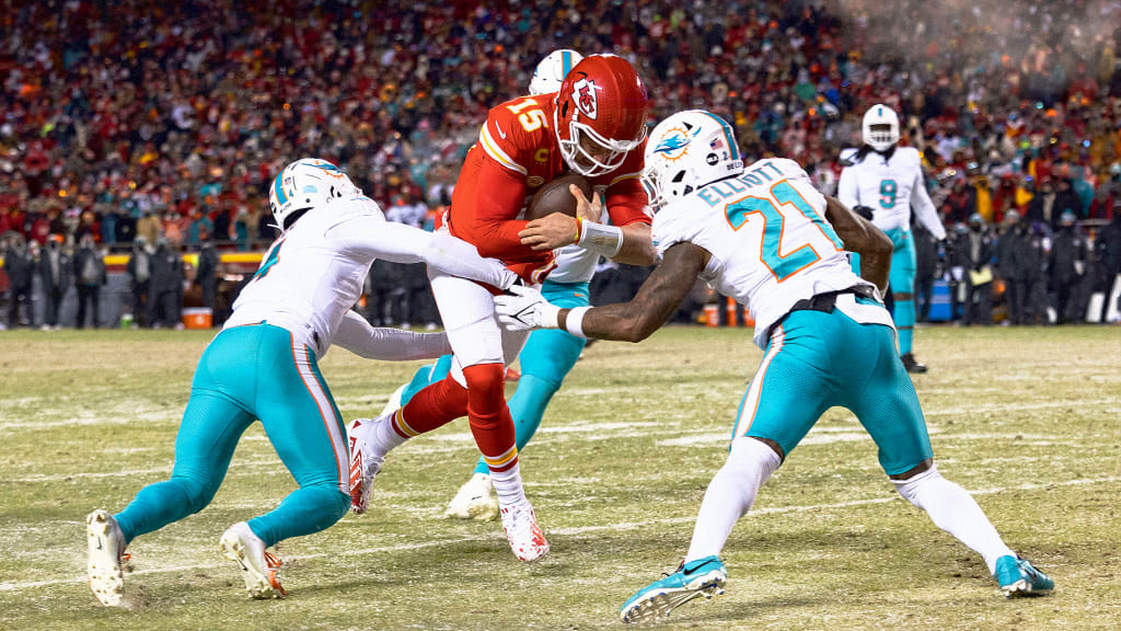 A football player in a red uniform and helmet tries to evade two opposing players in white and teal uniforms on a field. The background features a crowd of spectators under stadium lights.