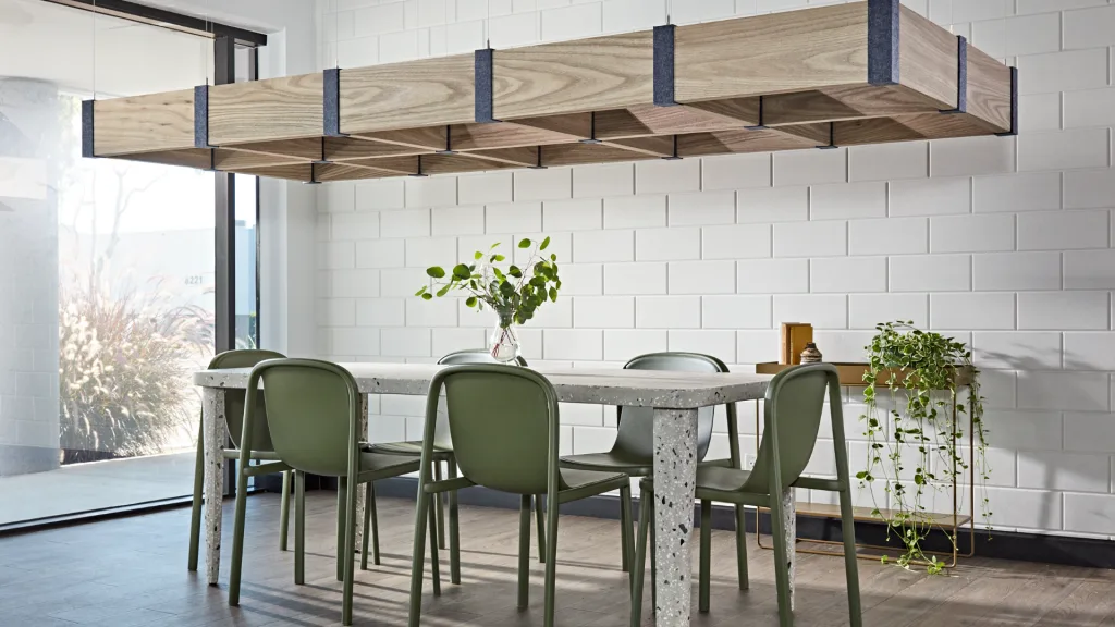 Modern dining area with a concrete table surrounded by green chairs. A large, rectangular wooden light fixture hangs above. The white tiled wall features small plants on the table and a stand. Sunlight illuminates the space through a glass door.