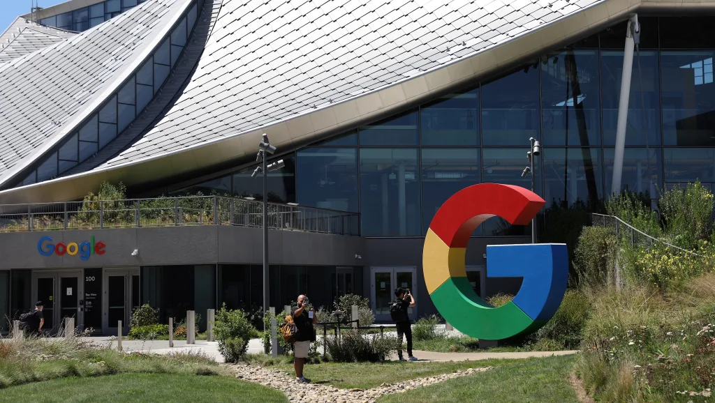 Exterior view of a building with large glass windows and a modern, wavy roof. In the foreground, theres a large, colorful Google G logo sculpture near well-maintained greenery. People are in front of the entrance doors.