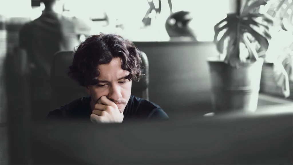 A person with curly hair is sitting at a desk, looking at a monitor with a thoughtful expression. The background is blurred, showing a large plant and a silhouette of another person walking by.