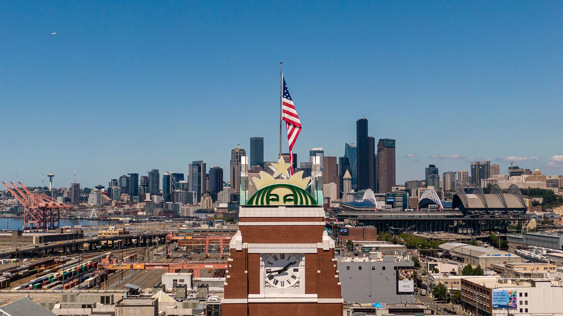 Aerial view of a clock tower with an American flag on top, set against the Seattle skyline. Skyscrapers rise in the background, including the iconic Space Needle on the left. The scene is under a clear blue sky.