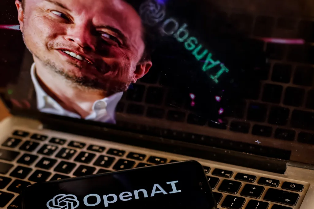 A laptop screen displays a distorted image of a persons face. A smartphone resting on the laptops keyboard shows the OpenAI logo with its text and emblem clearly visible.