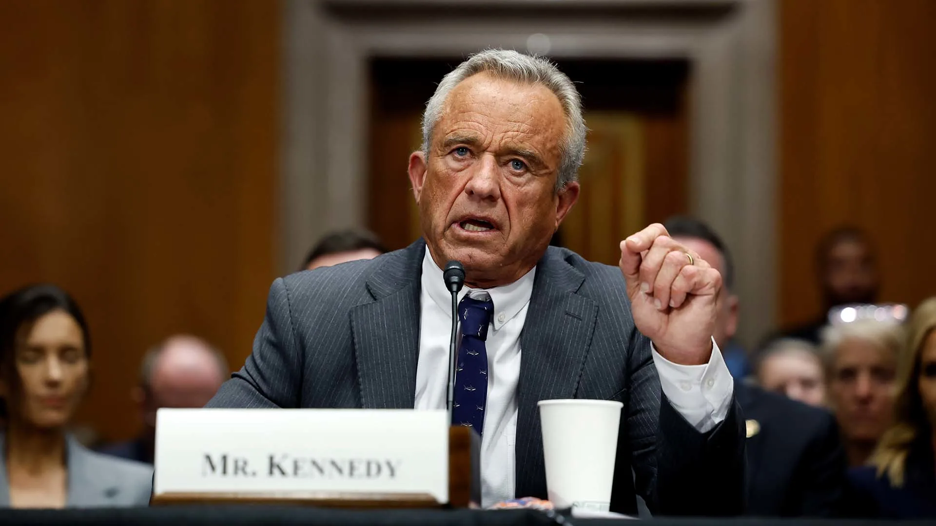 A man in a suit sits at a table speaking passionately, holding a pen. A nameplate reads Mr. Kennedy. A white cup is in front of him. Blurred people and a wood-paneled background are in the room.