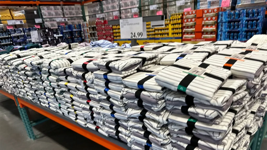 A large warehouse shelf filled with neatly stacked packs of white striped shirts, secured with different colored bands. Price sign above reads $24.99. Background shows shelves stocked with various products.