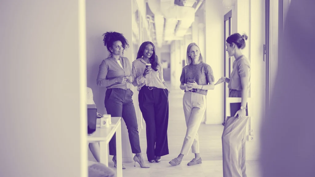 Four women gathered in a bright office hallway, holding coffee cups and engaging in conversation. The scene has a purple tint, and they are casually dressed, exuding a relaxed, professional atmosphere.