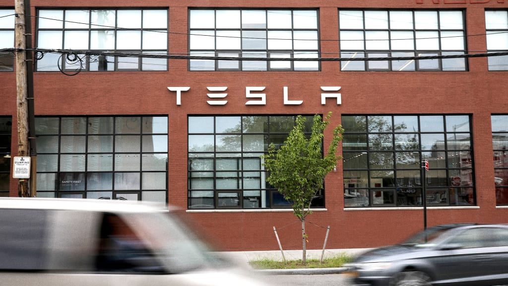 A brown brick building with large windows displays the Tesla logo. A blurred car passes in front on the road, and a small tree stands on the sidewalk.