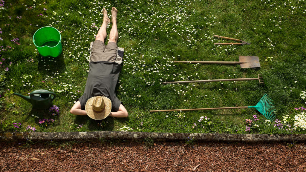 A person wearing a straw hat and shorts lies on their back on a grassy area filled with small white flowers. Garden tools like a rake, shovel, and hoe are neatly lined up nearby, along with a green bucket and watering can.