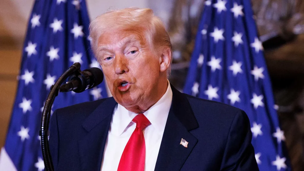 A man with light hair speaks into a microphone. He is wearing a dark suit, white shirt, and red tie. Behind him are two American flags with white stars on a blue field. The background is softly focused.