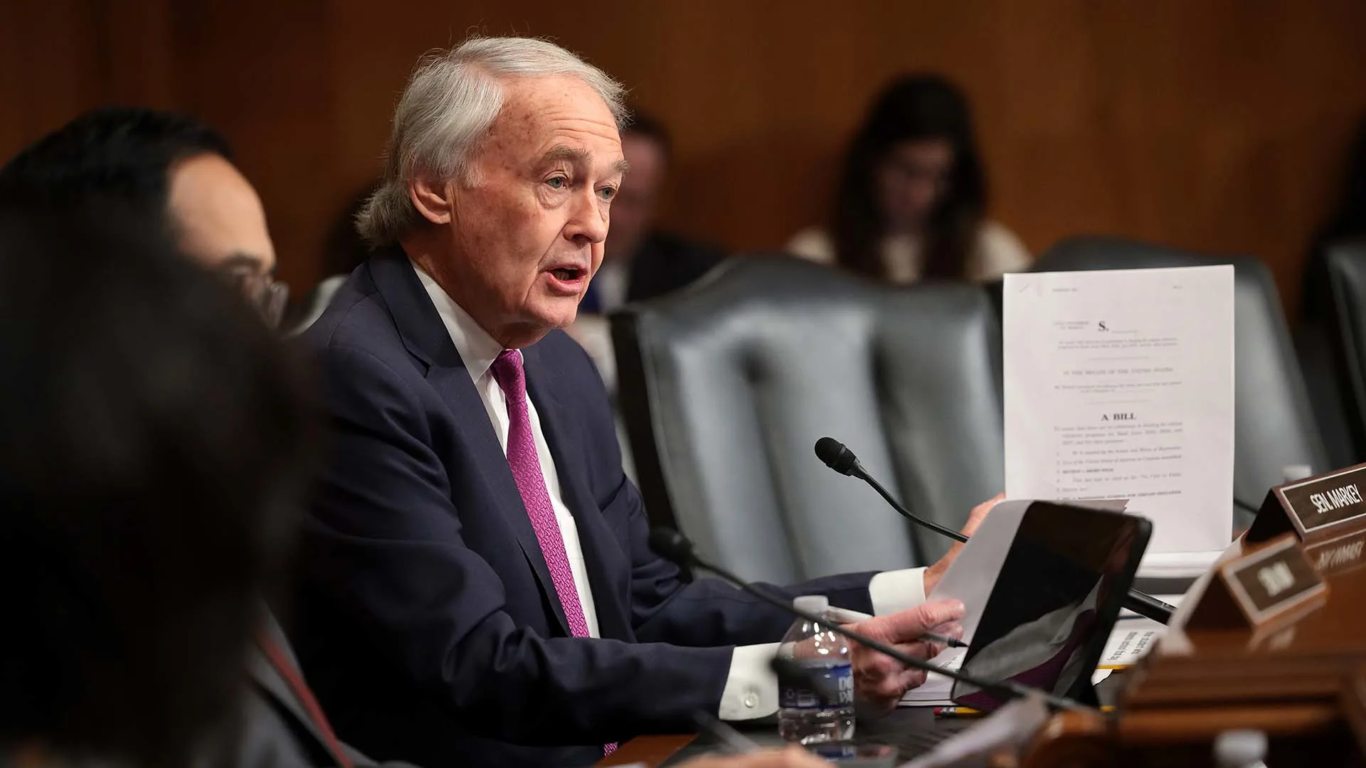 An older man in a suit sits at a table speaking, holding a document. Hes in a room with wood paneling and other people around him. Theres a microphone and a nameplate in front of him. People in the background are seated and looking attentive.