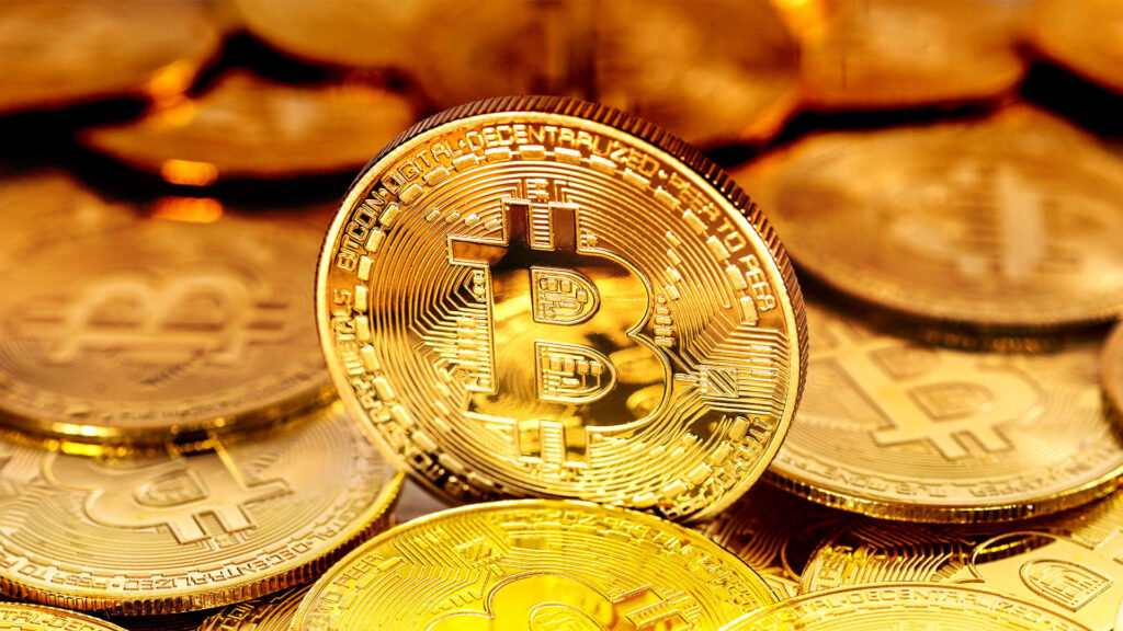 Close-up image of several gold coins with the Bitcoin symbol. The center coin is prominently tilted, displaying the Bitcoin B logo with circuit-like patterns. The background is filled with similar coins, creating a shimmering golden effect.