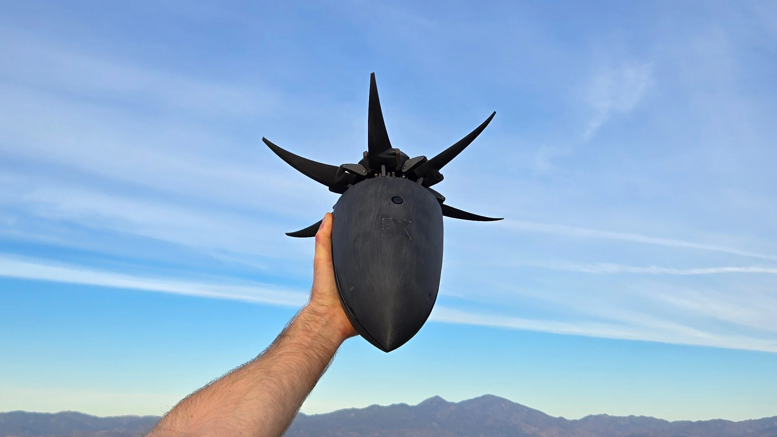 A hand holds a black, futuristic-looking drone with multiple upward-facing blades against a clear blue sky. Mountains are visible in the distant background.