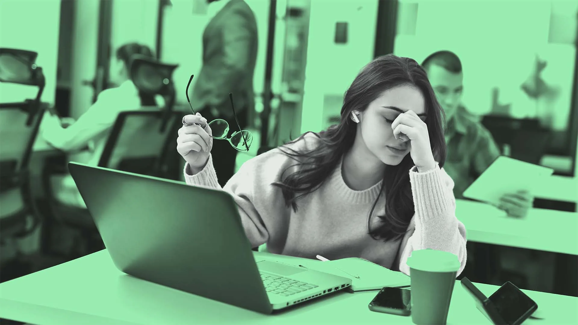 A woman sits at a desk with a laptop, holding her glasses in one hand and pinching the bridge of her nose as if stressed or tired. A coffee cup and smartphone are on the table. Other people work in the background.