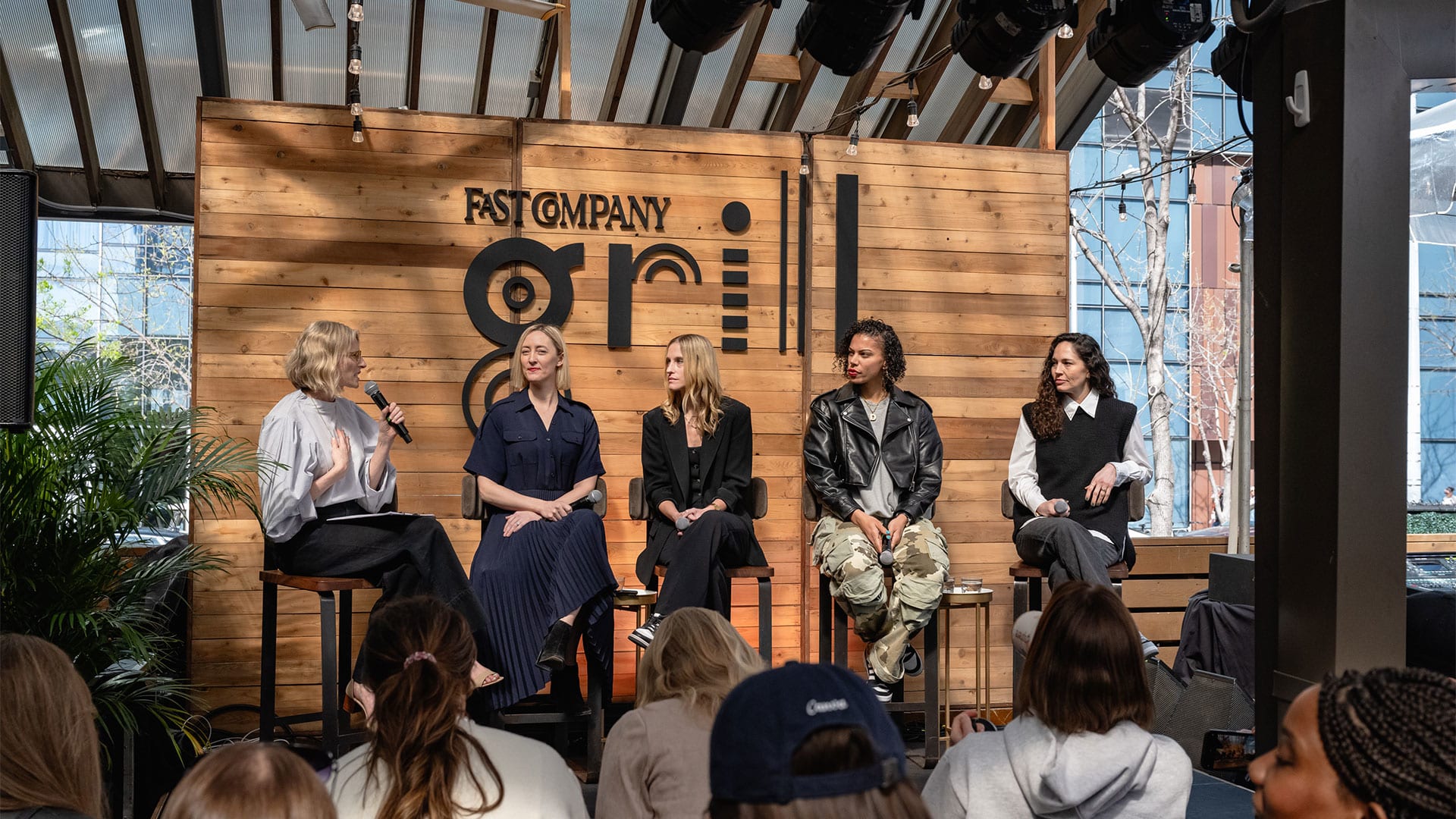 Five women sit on stools speaking on a panel at the Fast Company Grill event. An audience listens attentively in front of them. The background is wooden with the event logo displayed.