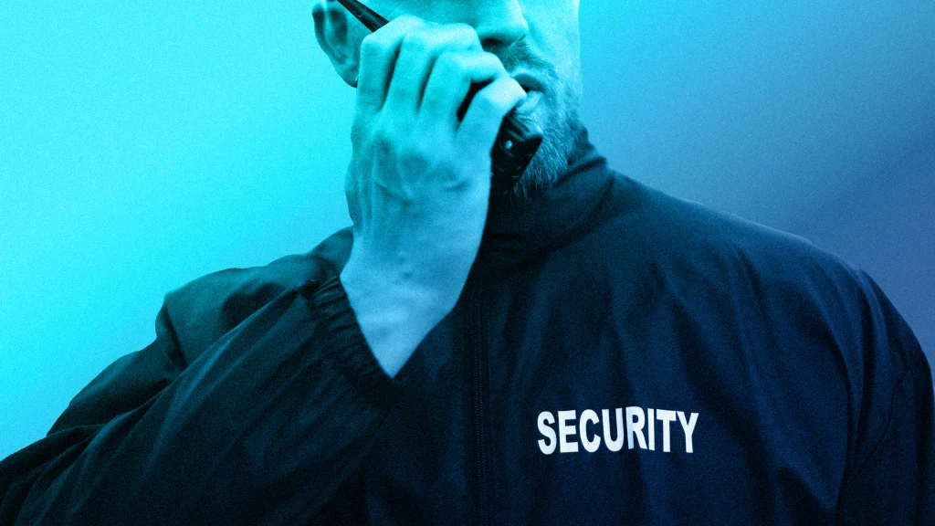 A person wearing a jacket labeled SECURITY holds a walkie-talkie near their mouth, appearing to communicate. The image uses a blue color filter.