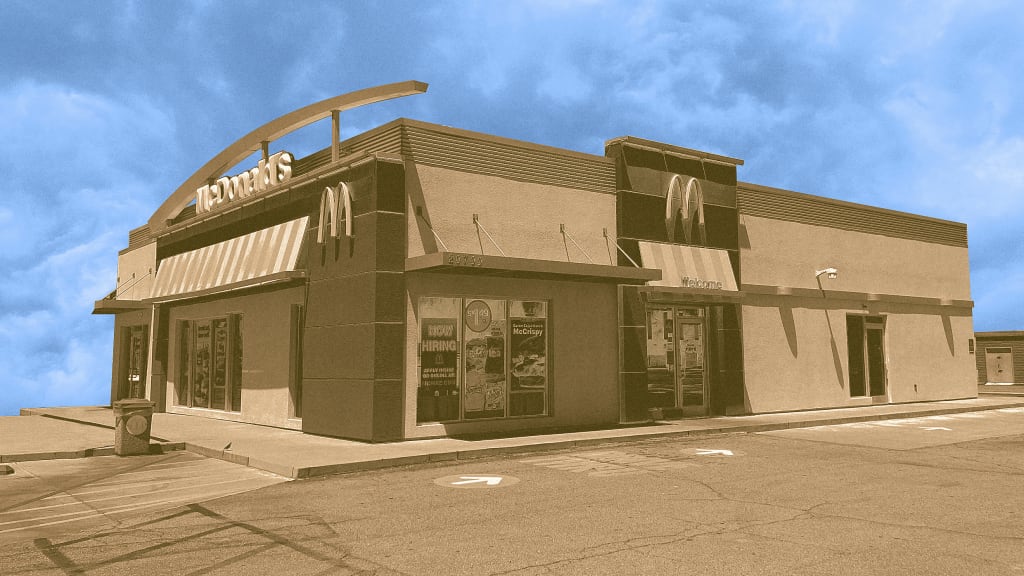 A sepia-toned image of a McDonald’s restaurant with signage, drive-thru, and promotional posters on the windows, set against a partly cloudy blue sky in an empty parking lot.