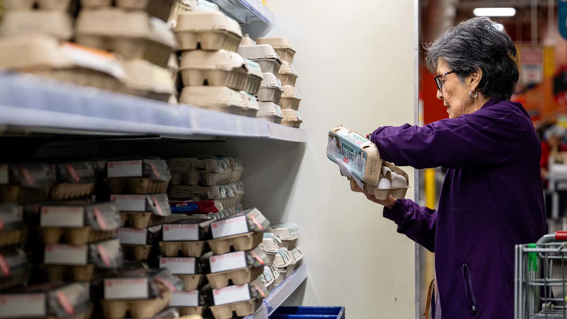 An older woman wearing glasses and a purple jacket selects a carton of eggs from a supermarket shelf filled with stacked egg cartons. She stands next to a shopping cart.