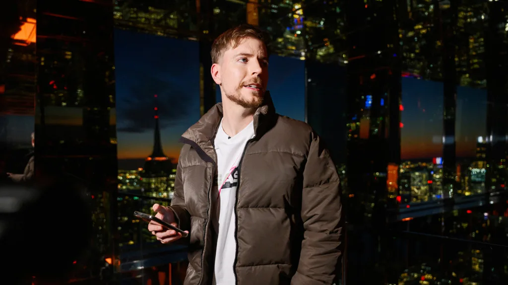 A man in a brown jacket holding a smartphone stands in front of large glass windows at night, with city lights and a dark blue sky in the background.