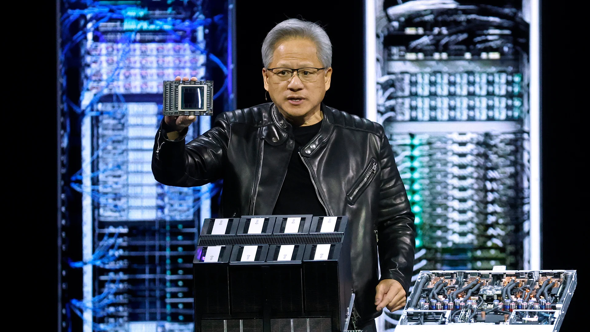 A man in a black leather jacket holds up a computer chip on stage in front of illuminated server racks, with various computer hardware displayed on the table beside him.