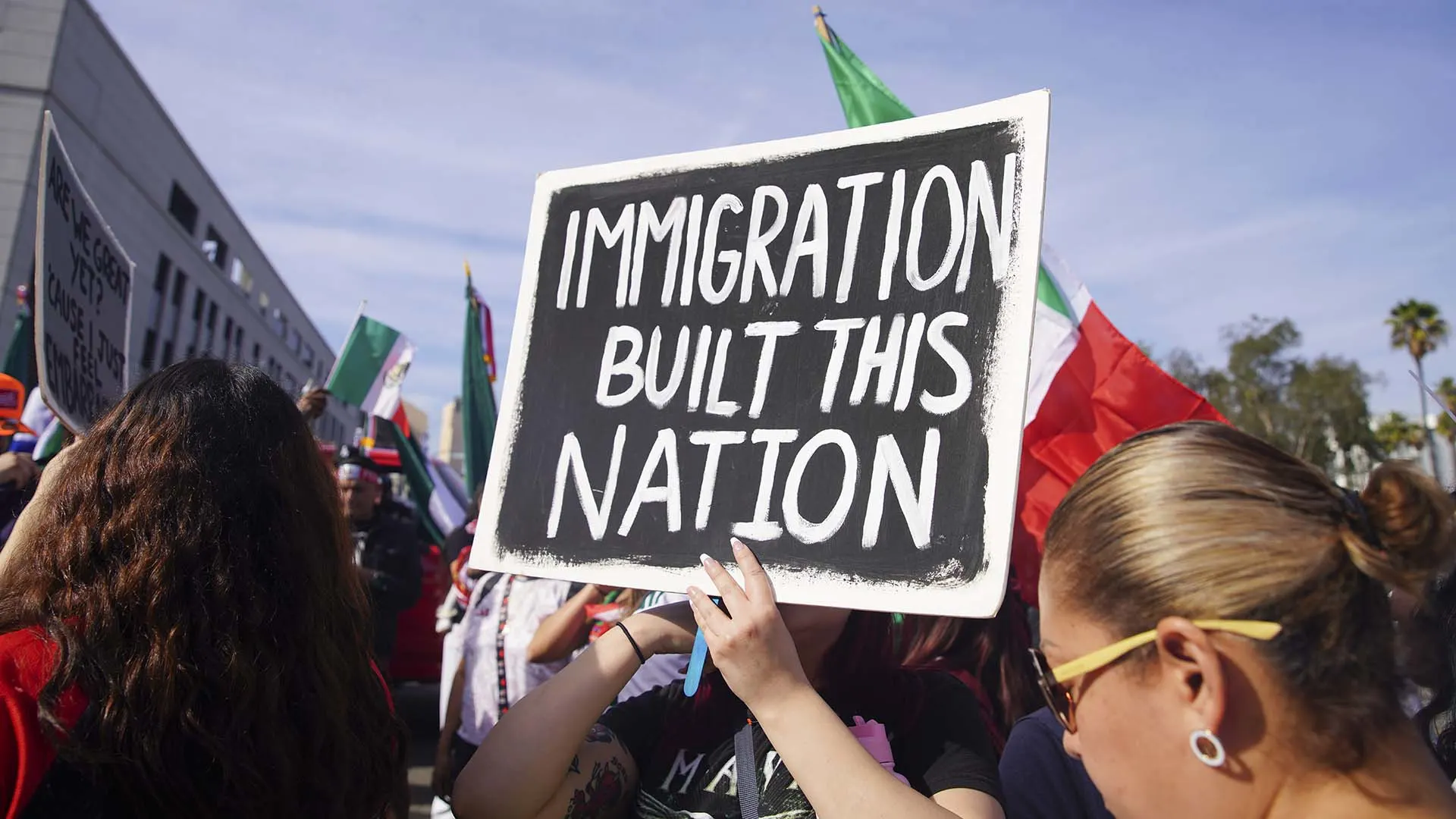 A person holds a large sign reading IMMIGRATION BUILT THIS NATION at a crowded outdoor rally, with flags and people visible in the background under a sunny sky.
