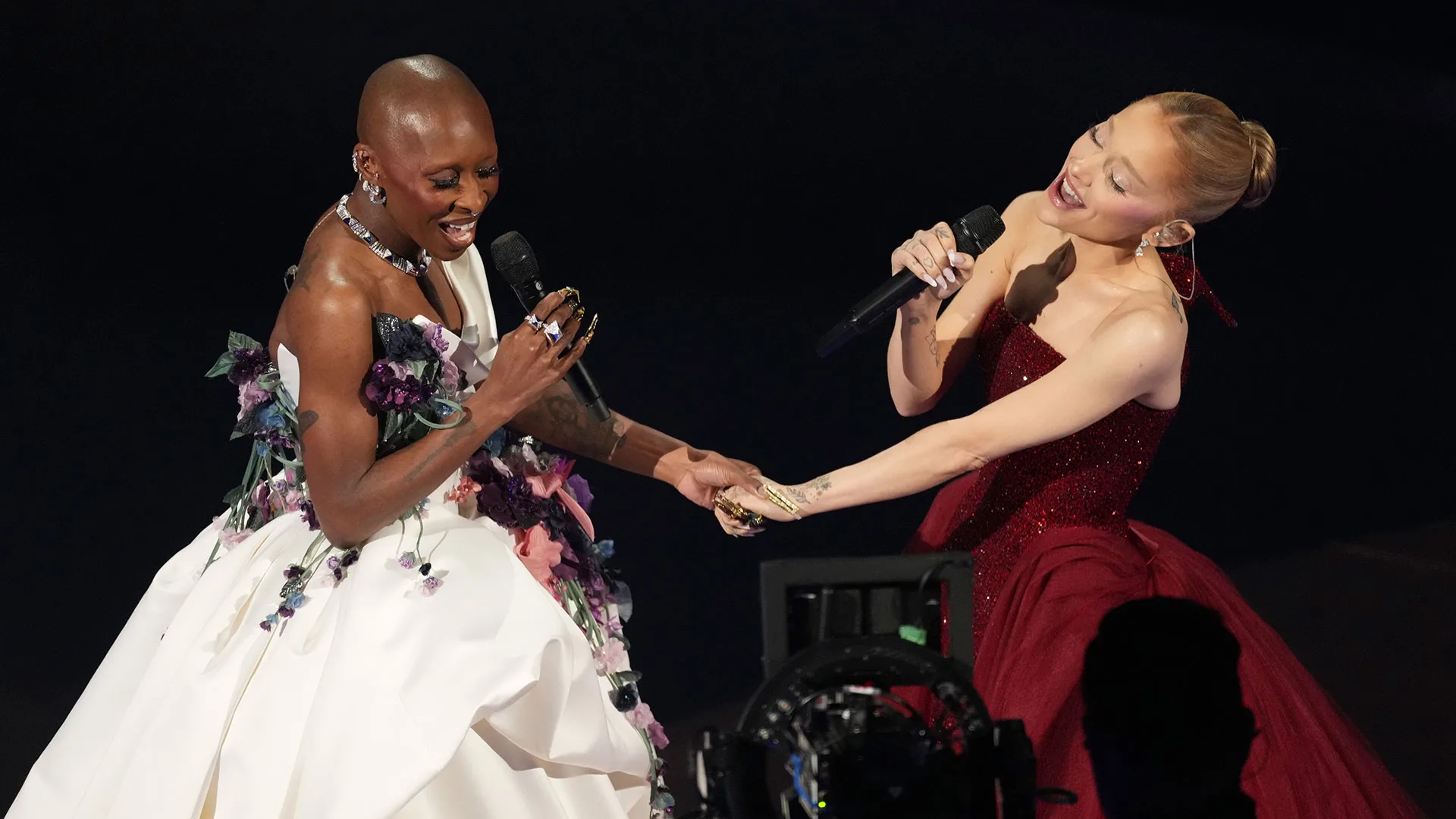 Two women in elegant gowns sing into microphones while holding hands on stage. One wears a white floral dress, and the other wears a red strapless gown. Both appear joyful and engaged in their performance.