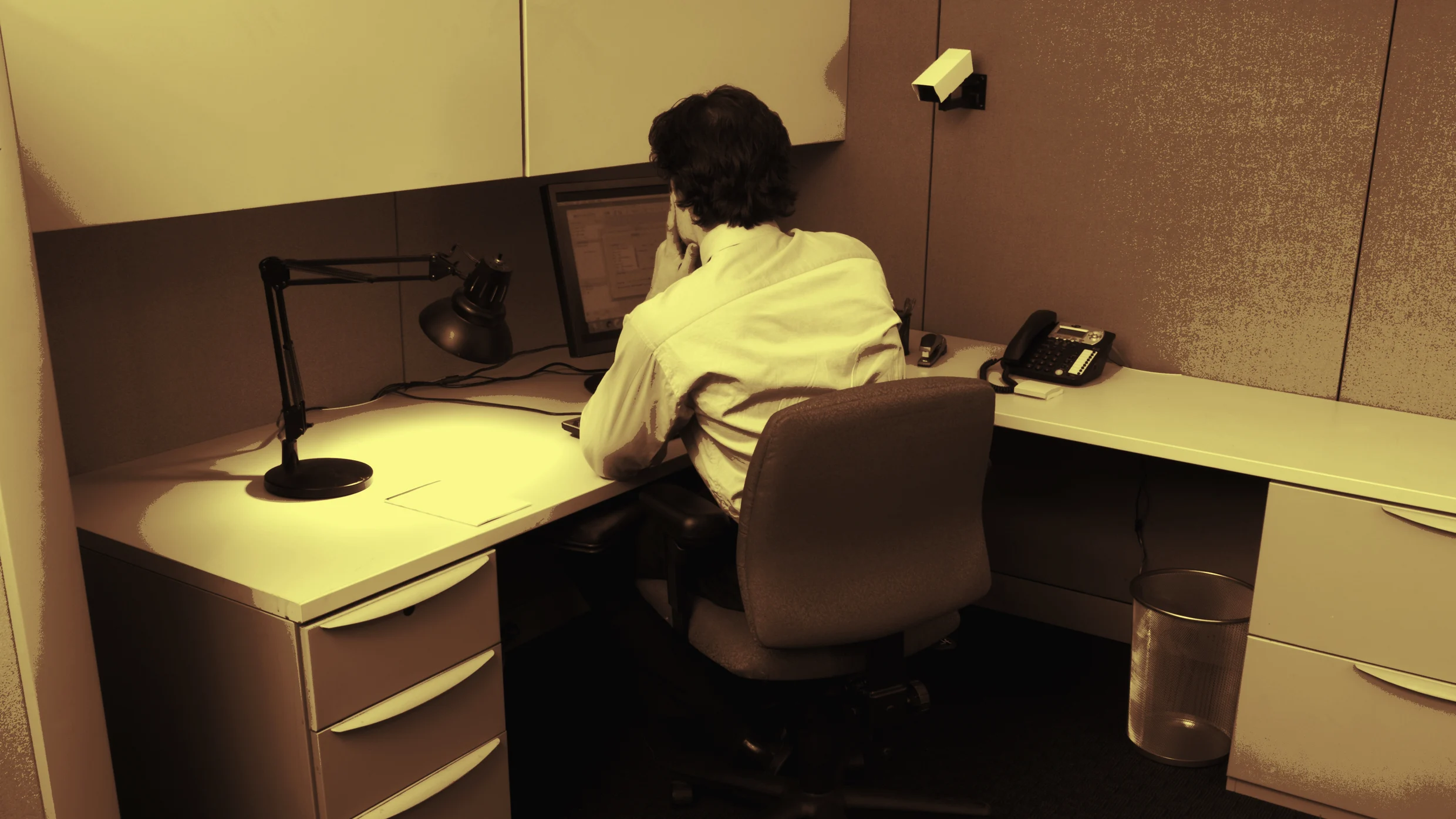 A man in a white shirt sits at a desk in an office cubicle, looking at a computer monitor. The workspace has a desk lamp, phone, and office supplies. The scene has a sepia tone.