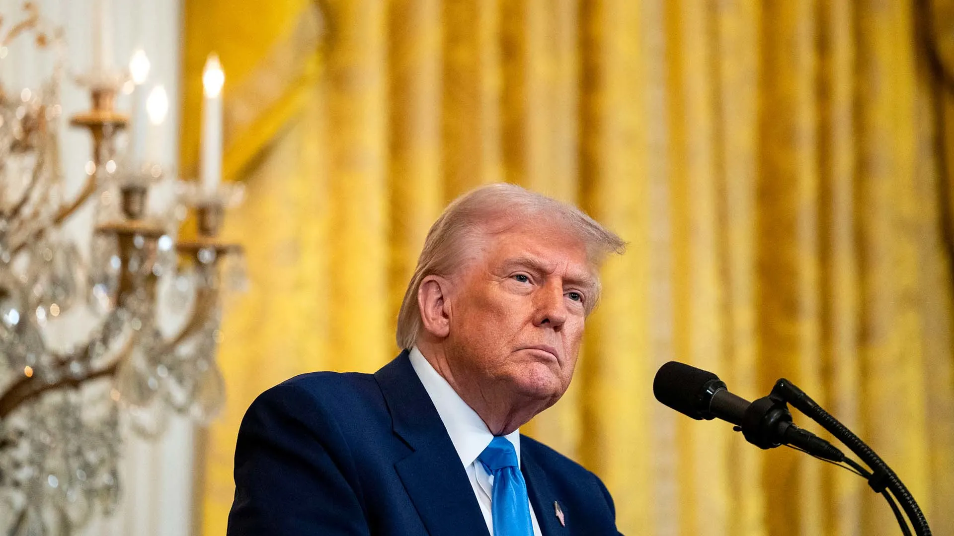 A man in a blue suit and tie stands at a podium with a microphone, looking serious. The background features golden curtains and a lit chandelier.
