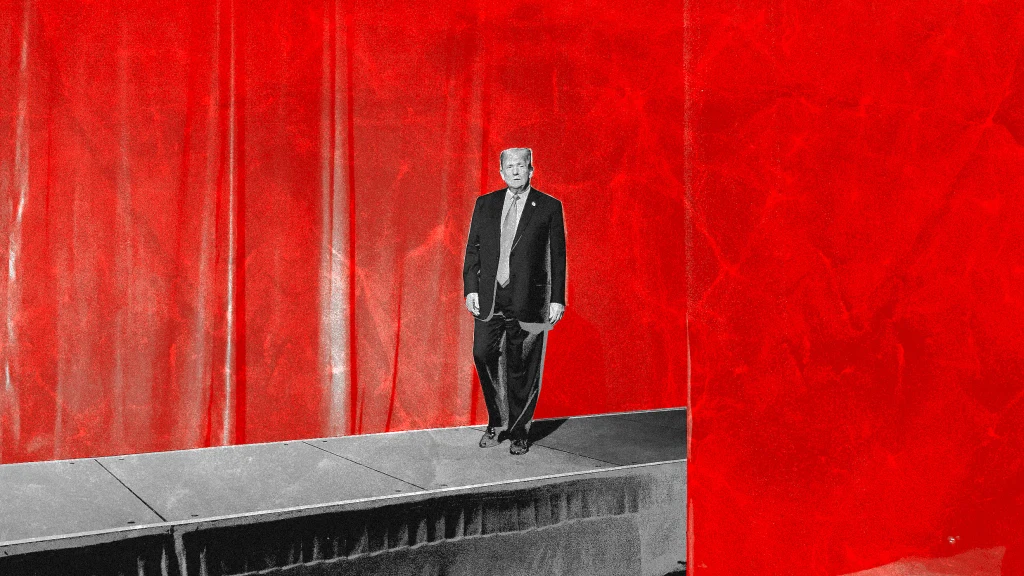 A man in a suit stands alone on a small stage against a large, bright red backdrop with dramatic lighting and shadows.