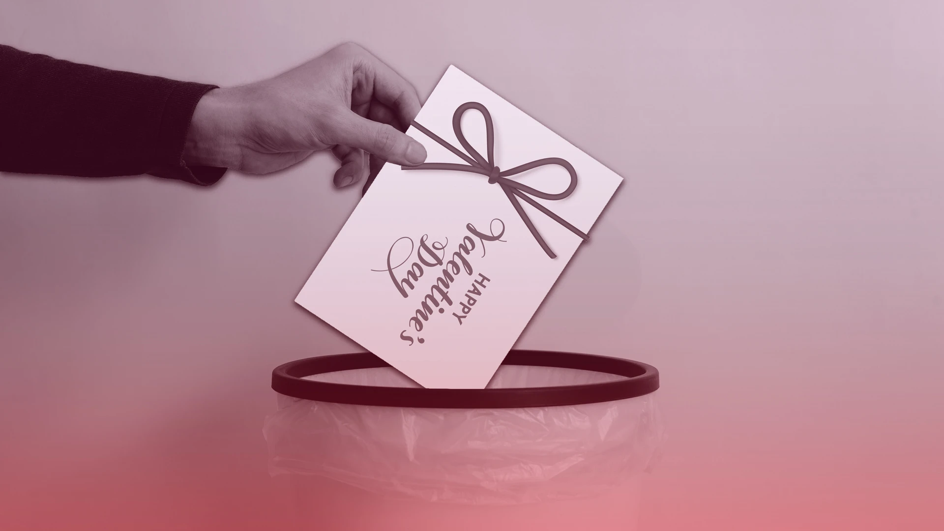 A hand drops a Happy Valentines Day card with a ribbon into a trash can lined with a plastic bag. The image has a pinkish tint.